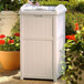 A beige Suncast outdoor waste container with a metal handle in a garden.