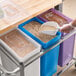 A person in a plastic glove using a scoop to put food into a white, blue, and purple Baker's Lane ingredient bin.