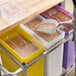 A person pouring grain into a white, yellow, and purple Baker's Lane ingredient bin.