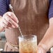 A person using a Barfly stainless steel 4-prong pineapple swizzle stick to stir a drink in a glass on a counter.