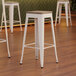 Three Lancaster Table & Seating white metal bar stools with gray wood seats.