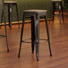 Three Lancaster Table & Seating black metal barstools with gray wood seats on a wood floor.
