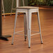 A Lancaster Table & Seating silver metal stool with a gray wood seat on a wood floor.