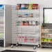 A Regency chrome mobile storage display stand with shelves of snacks and drinks in a convenience store.