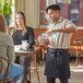 A man wearing a blue denim half bistro apron with black webbing pouring water into a glass on a counter.