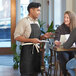 A man wearing a black Acopa Kennett bib apron talking to a woman at a farm-to-table restaurant table.