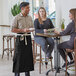 A man and woman sitting at a table with a man standing behind them. A man in a black Acopa Kennett Bistro Apron with natural webbing.