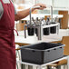 A man in a red apron holding a Choice stainless steel flatware carrier with black plastic containers over it.
