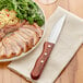 An Acopa steak knife on a napkin next to a plate of meat and vegetables.