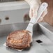 A person holding a Choice white square turner over a steak cooking in a pan.