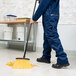 A person in blue overalls using a Carlisle heavy duty angled broom to sweep the floor in a professional kitchen.