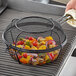 A hand using an Outset grill basket to cook vegetables on a grill.