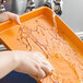 A person washing a Baker's Mark orange non-stick sheet tray on a counter with a sponge.