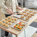 A woman using tongs to grab food from a Baker's Mark stainless steel sheet pan on a table.