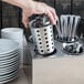 A hand picking up a container of stainless steel flatware cylinders filled with silverware.