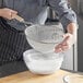 A woman using a Vollrath double mesh strainer with a wood handle to sift white powder into a bowl.