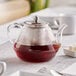 An Acopa glass teapot with brown liquid and a stainless steel infuser.