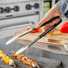 A person holding Vollrath stainless steel scalloped tongs over food on a grill.