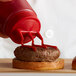 A person using a Vollrath red Tri Tip wide mouth bottle cap to pour ketchup on a burger.