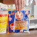 A person holding a Pop Weaver All-In-One Naks Pak Butter Burst Popcorn Kit bag in front of a popcorn machine.