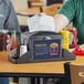 A condiment caddy on a table with a Choice napkin dispenser holding a napkin and ketchup bottles.