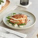 An Acopa Pangea porcelain plate with a piece of cooked chicken and a fork on a table.