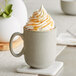 A close-up of a white Acopa Pangea mug filled with coffee and whipped cream with caramel sauce drizzled on top.