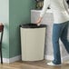 A person in jeans puts a bag in a beige Lavex corner round trash can.