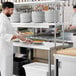 A chef in a white uniform plating food on a Regency expeditor table with a strip warmer.