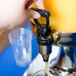 A hand using a black handle to pour juice from a Tablecraft juice dispenser into a plastic cup.