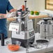 A person using an AvaMix commercial food processor to shred carrots in a stainless steel bowl.