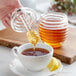 A hand using a Fox Run acrylic honey dipper to pour honey into a cup of tea.