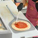 A square perforated aluminum pizza peel with a long handle being used to prepare a pizza with tomato sauce.
