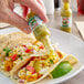 A person's hand pouring Marie Sharp's Green Habanero Hot Sauce onto a taco.