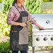 A woman wearing a black Backyard Pro grilling apron and holding tongs next to a grill.