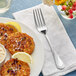 An Acopa stainless steel dinner fork on a napkin next to a plate of food with a glass of water.