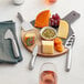 An Acopa stainless steel hard cheese knife set on a table with a cheese board with various types of cheese and fruits.