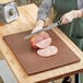 A person cutting meat on a Choice brown polyethylene cutting board.