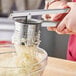 A person using a 10" stainless steel potato ricer to squeeze potatoes into a bowl.