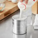 A person using a Choice aluminum measuring cup to pour milk.