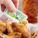 A hand holding a TABASCO® Green Hot Sauce portion packet over a plate of chicken wings.