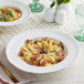 A plate of pasta with mushrooms and bacon in an Acopa Condesa porcelain pasta bowl on a table.