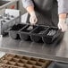 A person in a black apron holding a Choice black plastic cutlery box filled with utensils.