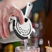A stainless steel Hawthorne strainer with four prongs being used to pour liquid into a glass filled with ice.