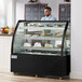 An Avantco refrigerated bakery display case with cakes on shelves and a man in a blue shirt behind it.