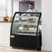 An Avantco black refrigerated bakery display case with cakes on shelves, a man in a blue shirt standing behind it.