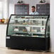 An Avantco refrigerated bakery display case with cakes on shelves behind a man in a blue shirt.