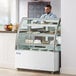 An Avantco white refrigerated bakery display case with a man in a blue shirt behind it.