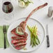 A Rastelli's bone-in tomahawk steak on a plate with asparagus and a glass of wine.