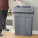 A man standing next to a Toter dark cool gray trash can with a black plastic bag inside.